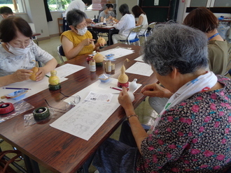 長机に並んで座った数名の女性たちが、手元にある図案や道具を使いながらひょうたんランプ作りを行っている写真