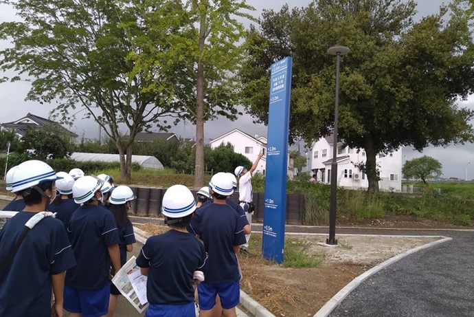 ヘルメットを被った多くの学生たちが、屋外で青い水位表示柱を見上げながら説明を聞いており、防災学習や地域の安全点検を行っている真剣な活動の様子を背後から捉えた、教育的な現場の風景を捉えた写真