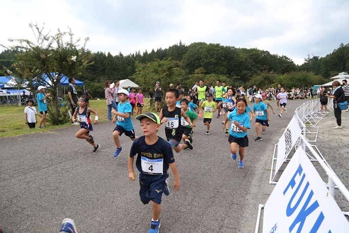 緑に囲まれた屋外のコースで、ゼッケンをつけた大勢の子供たちが一生懸命にゴールを目指して駆け抜けており、沿道から大人たちが温かい声援を送っている、活気あふれるマラソン大会のワンシーンを捉えた写真