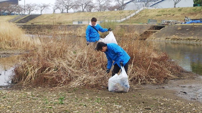 川辺の草むらで青い上着を着た二人がごみ袋を手に周囲の落ち葉やごみを丁寧に拾っている写真