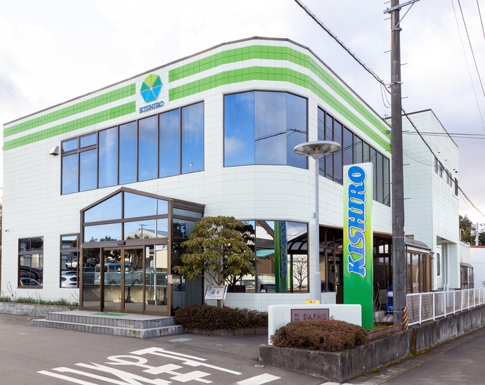 緑のグラデーションに「KISHIRO」と書かれた縦型看板が立った植栽の左側に、タイル調の白壁の上部にグリーンの2本の横線と7角形のロゴマークがあしらわれた、ガラス張りの大きな入口がある2階建ての建物がある写真