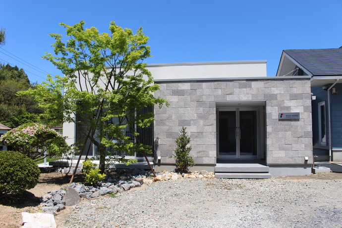 濃淡のグレーのブロック調の石壁で覆われた建物の中央にガラス戸の入り口が設けられ、砂利敷きの敷地の左側には植栽がある、株式会社熊田商店の外観写真