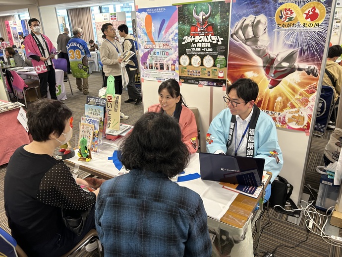 イベント会場のブースにてウルトラマンなどの3枚のポスターが貼られたパーティションを背に、水色の法被を着た女性と水色の法被を着た男性のスタッフがノートパソコンを広げながら、対面に座る二人の来場者へ笑顔で話をしている様子の写真