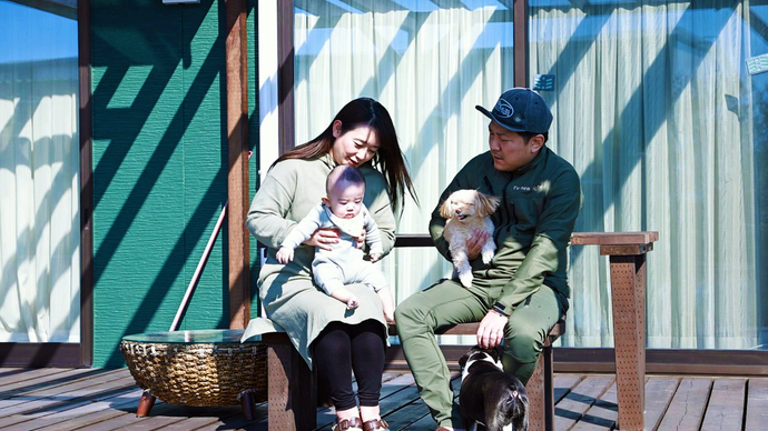 陽光が差し込むテラスのベンチに座り、増子さん夫婦が乳児と小型犬を抱えながら、足元にいる黒い犬を優しく見つめて微笑んでいる家族団らんの風景を写した写真