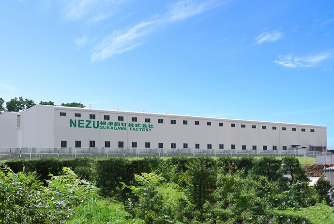 青空の下、白い平屋の工場または倉庫のような建物の壁に、緑色の大きな文字で「NEZU 根津鋼材株式会社 SUKAGAWA FACTORY」と書かれており、手前には緑の木々や低い生垣が広がっている写真