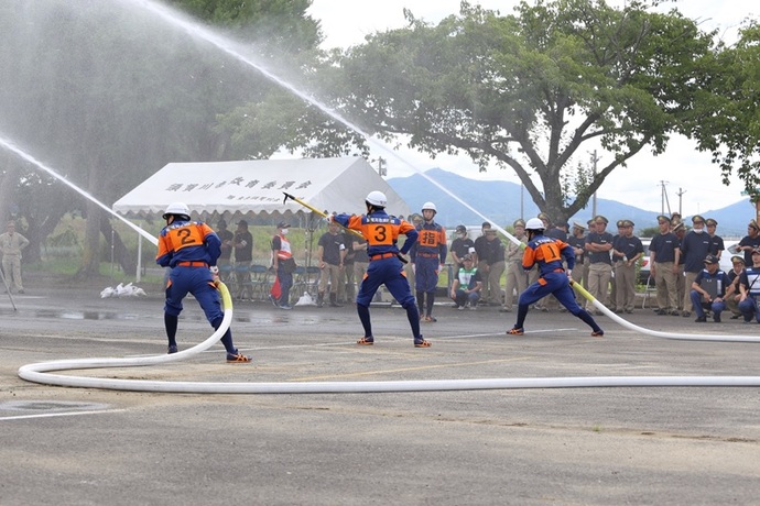 曇り空の下、オレンジと紺の訓練服を着用した消防団員たちが、白いホースを抱えて勢いよく放水作業を行っており、その様子を後方から多くの関係者が見守っている、緊迫感と規律正しさが伝わる消防演習の風景を捉えた写真