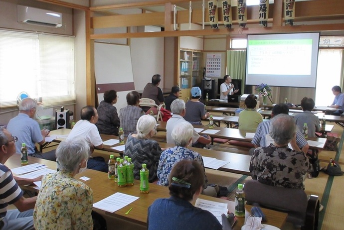 和室の研修室で、多くの高齢者が机に並んで座り、前方のスクリーンに映し出された資料を見ながら講師の話を熱心に聴いている、地域での講習会の風景を後ろから捉えた写真