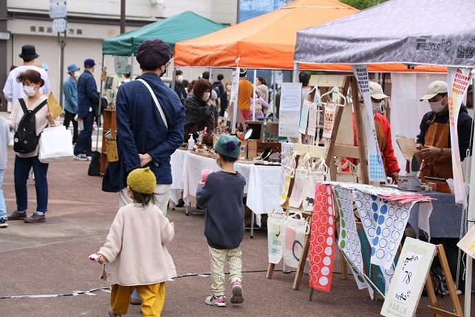 屋外の広場に色とりどりのテントが並び、手作りのバッグや小物が展示されたブースを親子連れや買い物客が楽しそうに巡っている、活気あるマルシェやフリーマーケットの賑やかな風景の写真