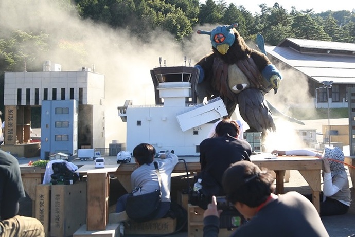 屋外の特撮セットで、巨大な怪獣の着ぐるみが煙の舞うビル群を破壊するシーンを、スタッフたちが低いアングルから真剣な表情で撮影している、迫力ある特撮映画の制作現場を収めた写真