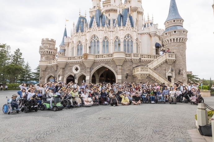 西洋の城を背景に、車椅子を利用する人々を含む多人数が広場に集まり、カメラに向かってポーズを決めたり笑顔を浮かべたりしている、賑やかで活気あふれる集合写真