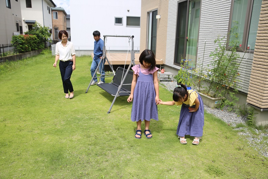 芝生が広がる明るい庭で、手前に立つ紫色のワンピースを着た二人の娘を、後方のブランコ付近から笑顔の両親が優しく見守っている写真