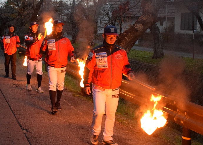 薄暗い中、赤いジャンバーと白いスラックスを身に着け、帽子をかぶった3名の選手と黒いスラックスの男性が火が点いた小松明を手にガードレール沿いを歩いている写真
