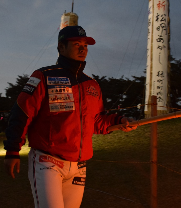 「祈 松明あかし」と書かれた大きな松明が2つ垂直にそびえたつ広場を背景に、法面下を歩く赤いジャンバーと白いスラックスを身に着け、帽子をかぶった一人の選手が、手に持った松明の火で赤く照らされている様子の写真