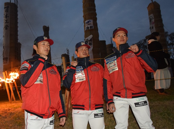 いくつもの大きな松明が垂直にそびえたつ広場を背景に、赤いジャンバーと白いスラックスを身に着け、帽子をかぶった3名の選手が片手で拳を作ったポーズをして並んで立っている写真