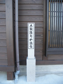 木造家屋の壁を背に、白い石造りの台座の上に「土蔵造りの商家」と縦書きで記された白い標柱が立っており、左側には雨どい、右側には格子窓が見える、歴史的建築物の詳細を伝えるための写真