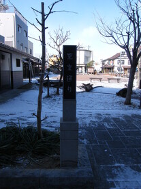 雪が薄っすらと積もった広場の入り口に、白い石造りの台座へ載った黒いプレートに縦書きで文字が記された石碑が立っており、背景には冬の青空の下、葉を落とした木々やベンチが点在する静かな公園のような風景が広がる写真