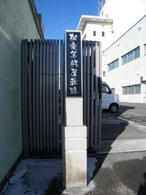 屋外の晴天の下、白い石造りの台座の上に「相楽等躬屋敷跡」と黒地に白の筆文字で記された縦長の石碑が立っており、背景には蛇腹式のゲートや白い建物、青空が広がる史跡の風景を捉えた写真