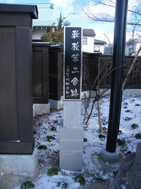 冬の晴天の下、白い石造りの台座に載った黒いプレートへ縦書きで文字が刻まれた石碑が立っており、周囲には雪が残る地面や黒い板塀、細い若木が配置された歴史的な情緒漂う史跡の風景を捉えた写真