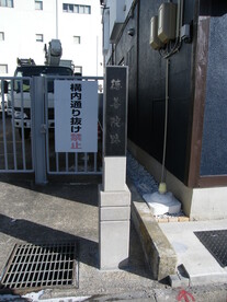 建物の脇に、白い石造りの台座に載った黒いプレートへ「徳善院跡」と縦書きで刻まれた石碑が立っており、背後には「構内通り抜け禁止」の看板が掛かった門扉や白い車両、暗い色の外壁の建物が見える、街中の史跡を捉えた写真