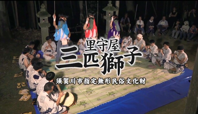 夜の静寂の中で、青いシートと茣蓙を敷いた舞台を囲み、浴衣姿の演奏者たちが奏でる音に合わせて、獅子頭を被り色鮮やかな装束を纏った三人の踊り手が舞を披露している「里守屋三匹獅子」の奉納行事を捉えた写真