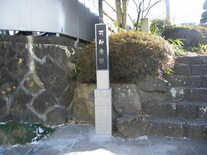 石垣と植栽を背に、白い石造りの台座に載った黒いプレートに白の筆文字で刻まれた縦長の石碑が立っており、右側には石段が続き、左側には銀色のフェンスが見える、歴史的な趣を感じさせる屋外の風景を写した写真