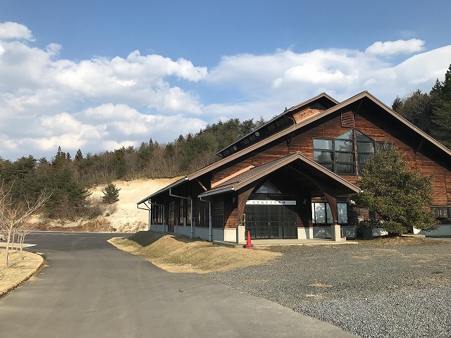 森を背景に、舗装された道の横にある砂利敷きの敷地に建つ、大きな三角屋根が特徴的な木造の建物の写真