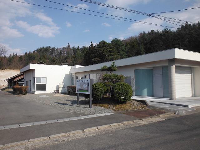 舗装された敷地に建つ、手前に案内板や植栽が配置された、白い外壁の平屋建ての施設の写真