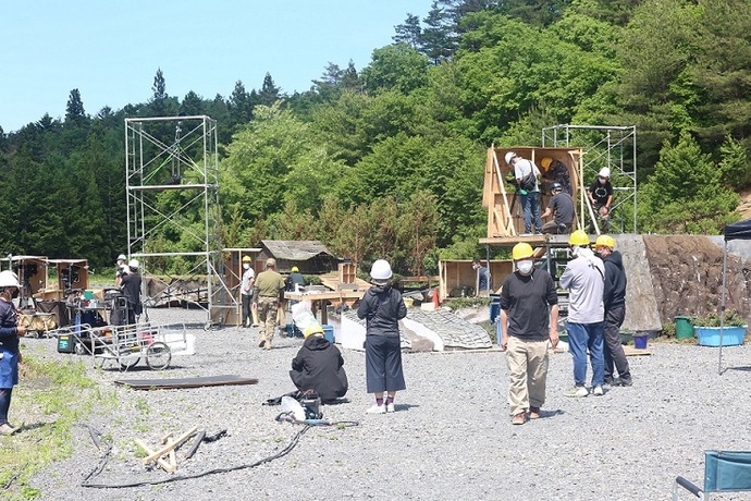 屋外のロケ現場で、ヘルメットやマスクを着用した多数のスタッフが緑豊かな森を背景に、足場や木製のセットの組み立て、機材の準備などの作業に取り組んでいる撮影風景の写真