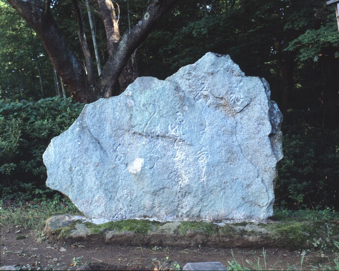 緑豊かな木々を背景に、表面に崩し字の和歌や俳句のような文字が深く刻まれた、ごつごつとした青みがかった自然石の石碑が、苔の生えた土台の上にどっしりと安置されている写真