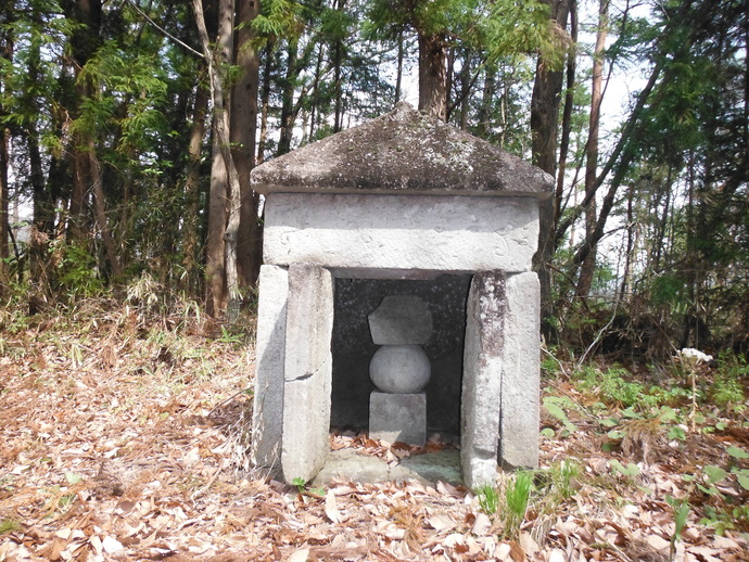 雑木林を背景に、石を積み上げて造られた小さな祠の中に、四角い台座の上に丸い石が乗った五輪塔が祀られている写真