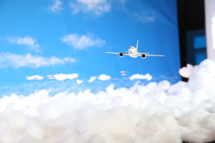 青い空と白い雲を背景に、綿で表現されたような真っ白でふわふわとした雲海の上を、正面を向いた小さな旅客機の模型が軽やかに飛行している様子をミニチュアで再現した写真