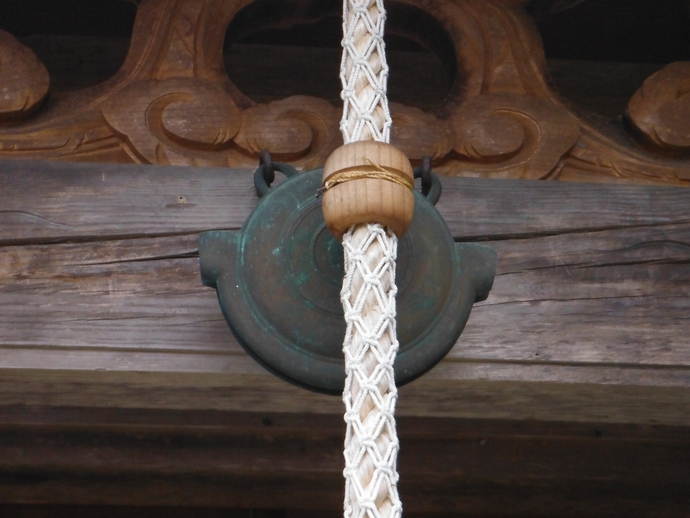 木製の梁に取り付けられた青銅製の鰐口が正面から写り、その手前に、木製の輪が通された白い編み込みの太い縄が垂れ下がっている写真