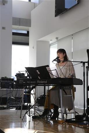 椅子に座って、楽譜を乗せたキーボード演奏しながら、マイクで歌唱している岡本真夜さんの写真