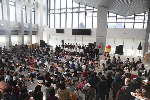 天井が高いホールの椅子やスタンド席に大勢の観客が集まり、前方のステージで岡本真夜さんとたくさんの児童・生徒が整列して合唱し、その横では赤い炎をモチーフにしたキャラクターボータンが両手をあげて盛り上げている様子を上から撮影した写真