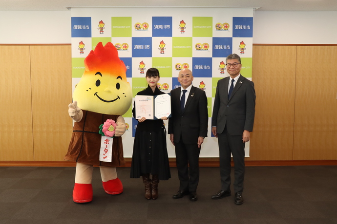 須賀川市のバックパネルの前で、マスコットキャラクターのボータンと、表彰状を手にする峰平 朔良(みねひら さくら)さん、スーツ姿の大寺市長と男性が並び、記念撮影を行っている写真