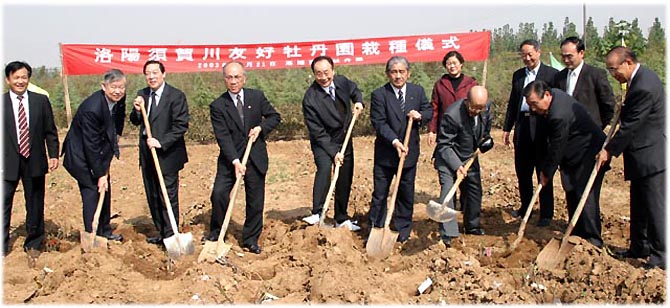 緑の森を背景に、赤に白文字で式典の横断幕が設置された茶色の土の土地に並んで立った8人のスーツ姿の男性が、手にしたシャベルで土を掘り返そうとしている、友好都市締結10周年記念植樹の際の記念写真