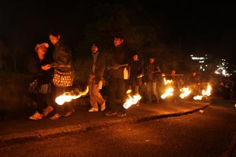 暗い夜道を火が点いた小松明を手に下げた人々が、列になって歩いている様子の写真
