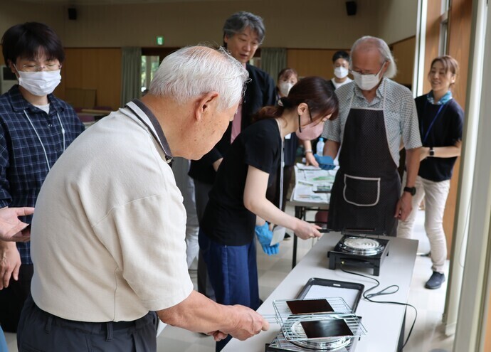 室内の明るいスペースで、テーブルの上に電気コンロと金網が並び、複数人が防蝕剤の固定をしている様子を写した写真