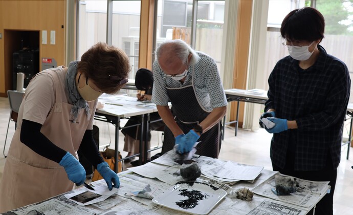 室内のテーブルに新聞紙が敷かれ、複数人が青い手袋を着けてインクつめの作業をしている様子を写した写真