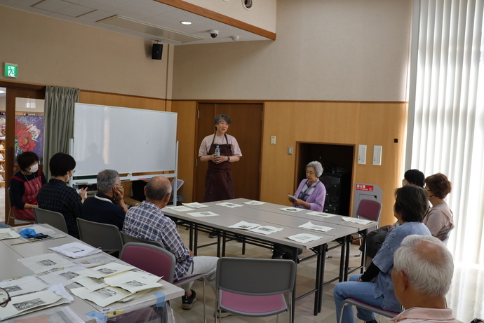 明るい室内で長方形のテーブルを囲んで複数人が座り、前方のホワイトボード近くでエプロンを着けた人物が説明をしている様子を写した写真