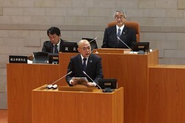 議場で、壇上に立った大寺市長が資料を手に持ち発言しており、後ろの議長席とその左側の席に、スーツ姿の男性が着席している、市議会定例会本会議の様子の写真