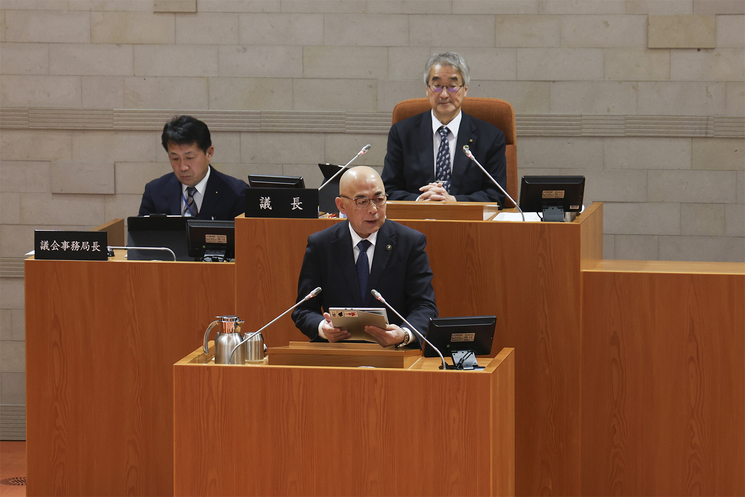 議会場のような場所で、スーツ姿の男性が演台に立ちタブレット端末を操作しながら発言しており、その後ろの席には別の男性たちが座って様子を見守っている、厳粛な会議や議会の進行風景を捉えた写真