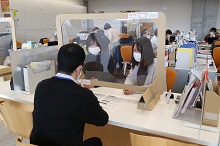 オフィスの受付カウンターで、飛沫防止用の透明なパーティション越しに、マスクを着用した職員と来客が対面して相談や手続きを行っている写真