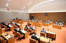 天井が高くアーチ状になった議場で、木製のデスクに座った大勢の議員たちが前方の議長席に向かって整然と並び、真剣な面持ちで審議に臨んでいる様子を後方から俯瞰で捉えた写真