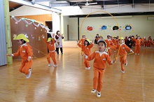 広々とした屋内のホールで、お揃いのオレンジ色のトレーニングウェアと黄色い帽子を身につけた園児たちが、一斉に縄跳びの練習に励んでいる写真