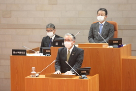 議場の演壇でスーツ姿の人物がマスクを着けて発言しており、背後には木製の机に座る議会関係者と書類が並ぶ市議会定例会本会議の写真