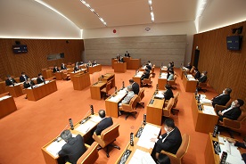広い議場で複数の人物が木製の机に着席し、中央に演壇がある配置で会議が進行している市議会定例会本会議の様子が写された写真