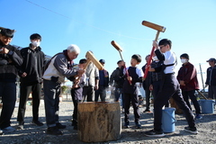 屋外で複数の人々が集まり、中央で木の臼を囲んで杵を振り上げ餅つきをしている様子が写された写真