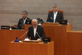 演壇に立ち答弁を行うスーツ姿の男性と、その後方の席に座る議長などと思われる二人の男性が議場に並び、厳かな雰囲気の中で会議が進行している様子を捉えた写真