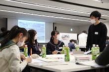 明るい会議室で、マスクを着用した制服姿の女子生徒たちと男性がテーブルを囲み、資料を広げて熱心に意見を交わしている、若者による地域課題解決やワークショップの様子を捉えた写真